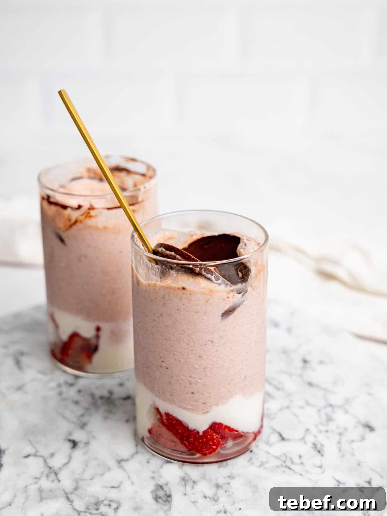 Two glasses of layered strawberry overnight oats on a marble surface, with strawberries and yogurt at the base, a creamy pink blended oat layer, and a chocolate topping, one with a spoon inserted.