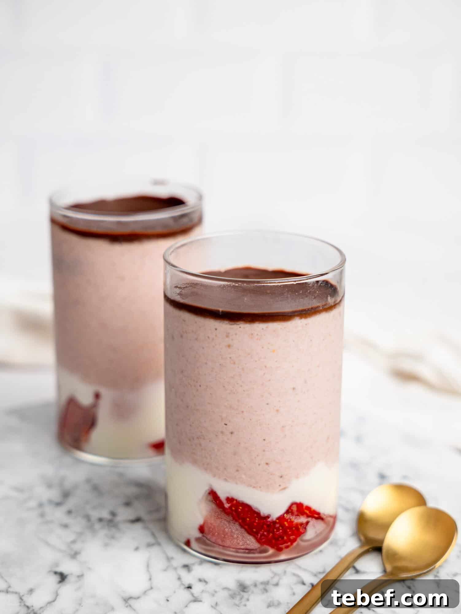 Two glasses of strawberry blended overnight oats on a marble surface with a smooth chocolate topping, with gold spoons placed beside them.