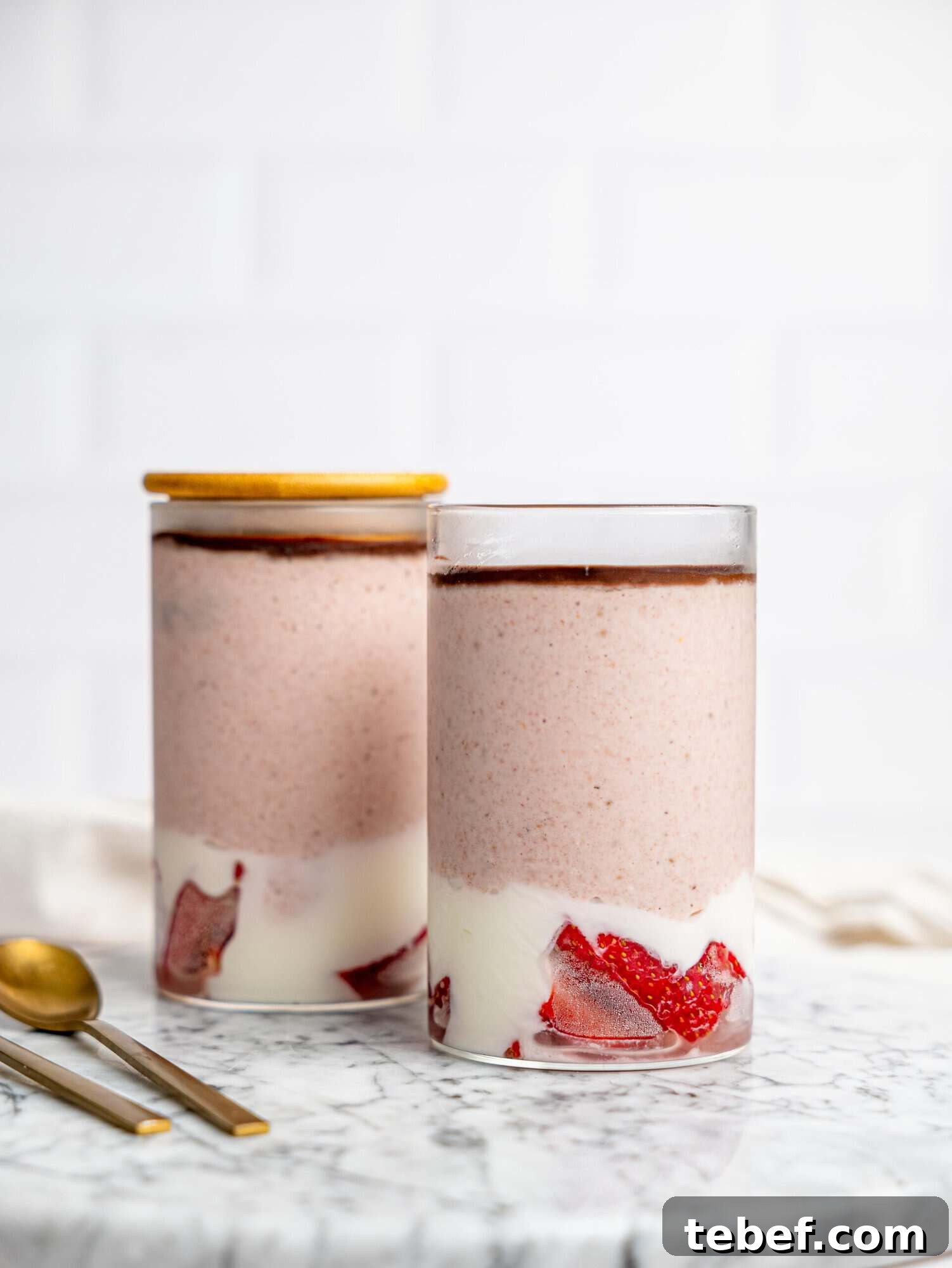 Two glasses of layered strawberry overnight oats on a marble surface, with strawberries and yogurt at the base, a creamy pink blended oat layer, and a thin chocolate topping, with spoons beside them.
