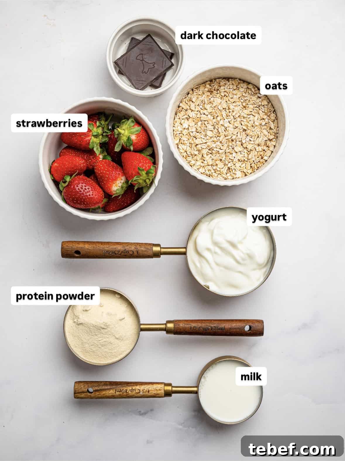 Flat lay of ingredients for blended overnight oats on a light surface, including fresh strawberries in a bowl, rolled oats, dark chocolate squares, yogurt, protein powder, and milk measured in wooden-handled cups.