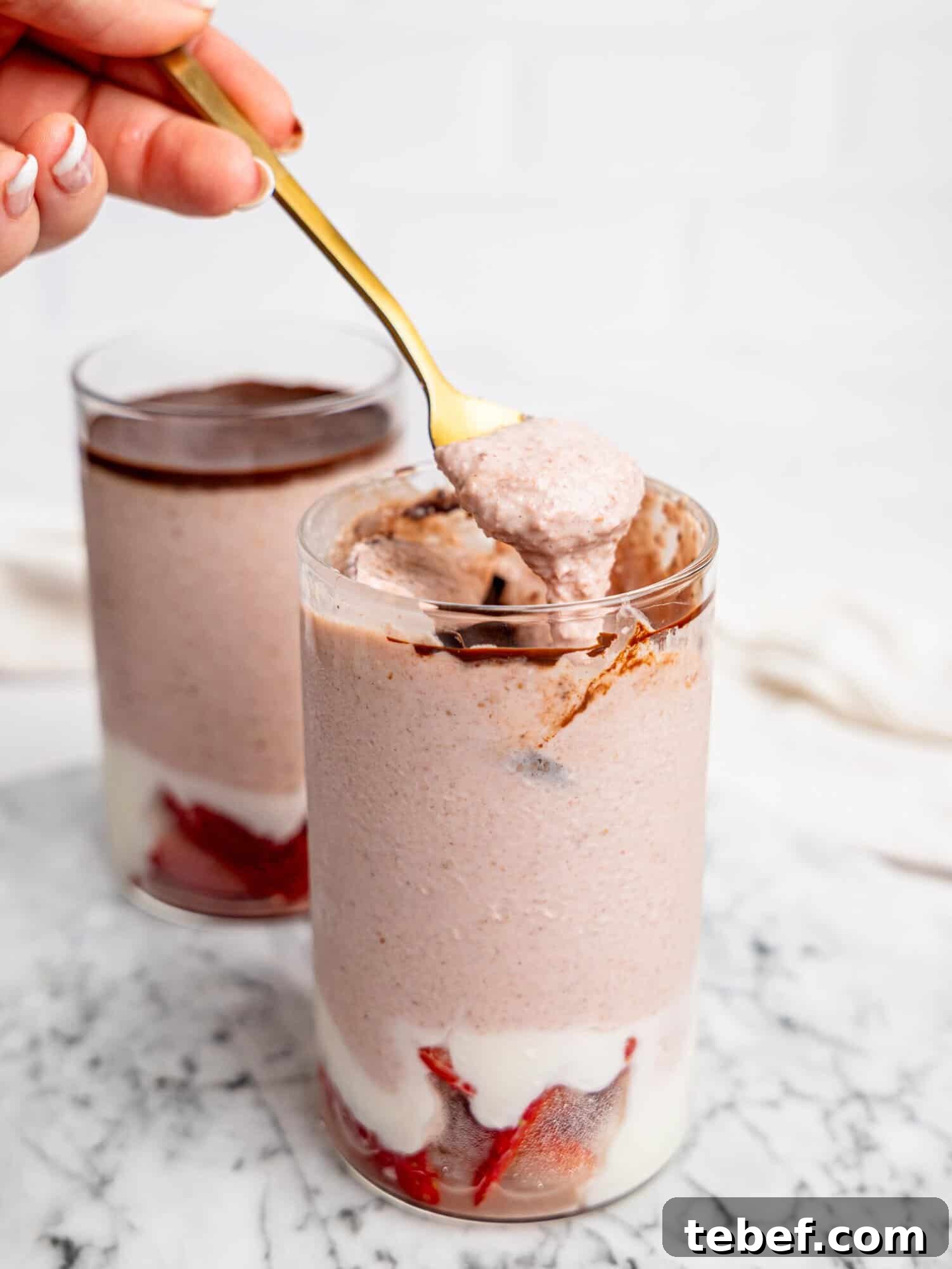 Close-up of a spoon lifting a creamy bite of strawberry blended overnight oats from a glass with visible chocolate layer and strawberry yogurt base.