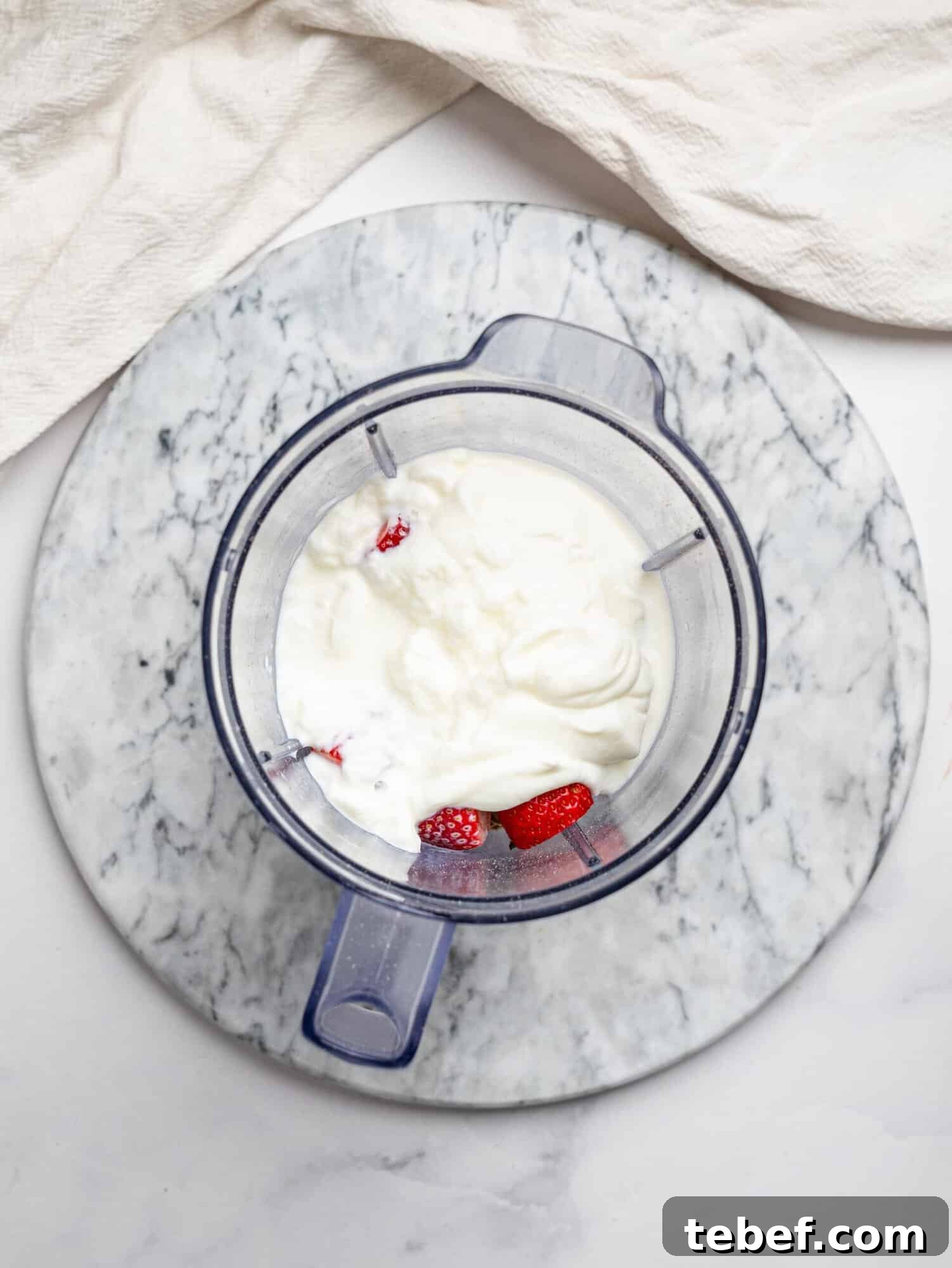 Top view of a blender jug on a marble board filled with ingredients for strawberry overnight oats, including yogurt and strawberries on top, with the remaining ingredients underneath ready to be blended.
