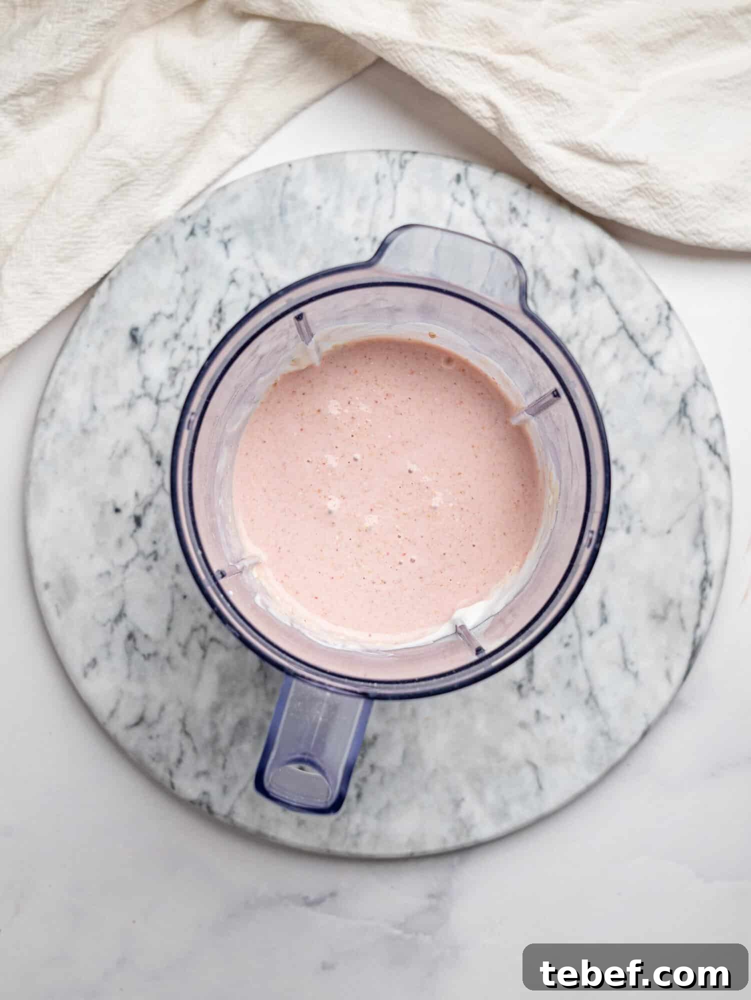Top view of a blender jug on a marble board filled with smooth, pale pink blended mixture for strawberry overnight oats.