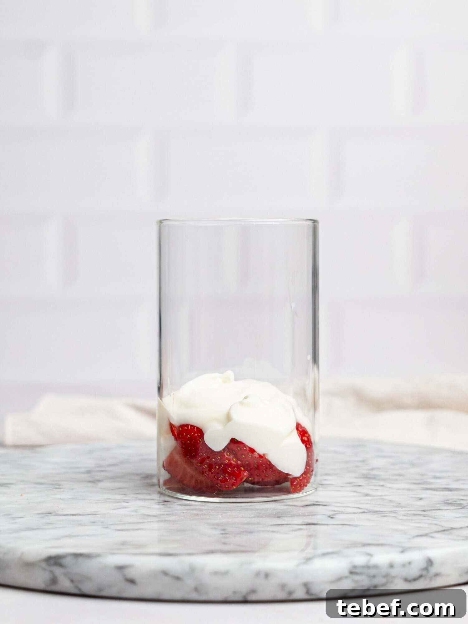 Side view of a clear glass on a marble surface with sliced strawberries and a dollop of yogurt at the bottom, ready for layered strawberry overnight oats.