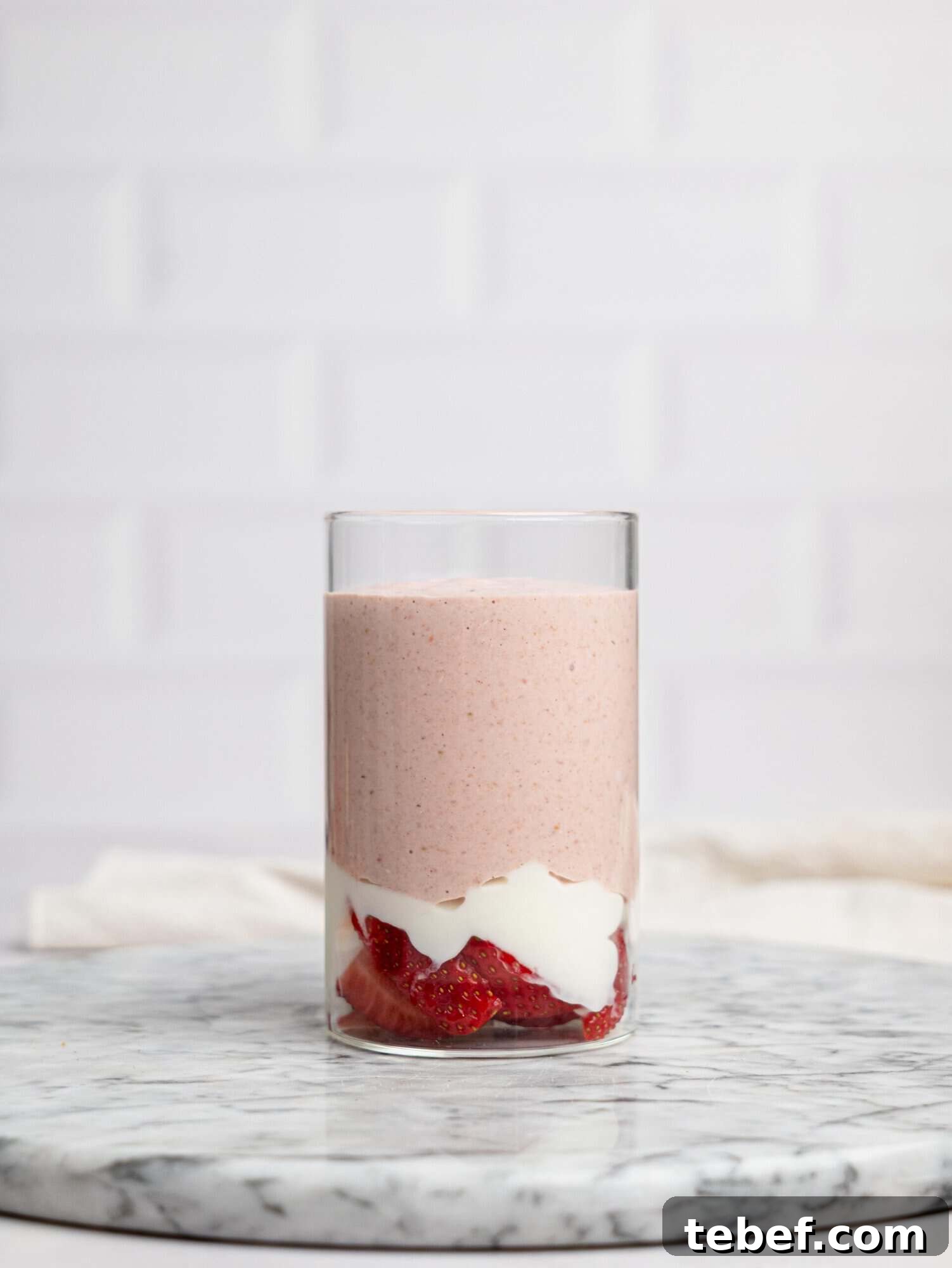 Side view of a clear glass on a marble surface layered with sliced strawberries, yogurt, and a smooth pink blended mixture for strawberry overnight oats.