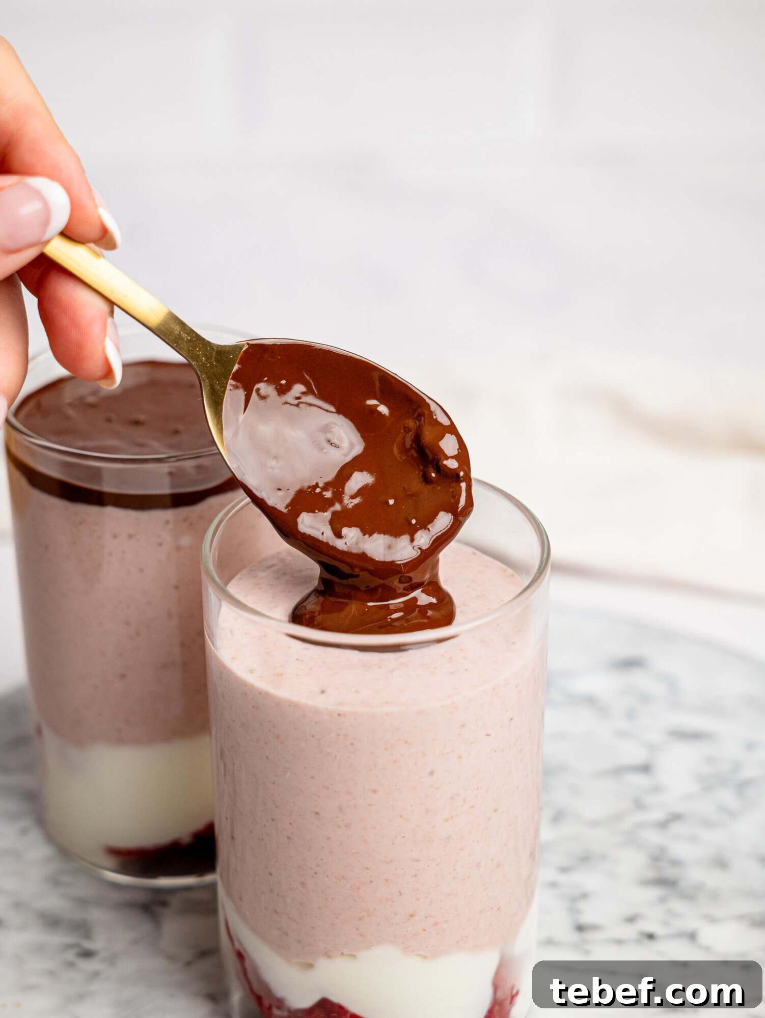 Close-up of a hand spooning melted chocolate over a glass of layered strawberry overnight oats with strawberries, and yogurt.