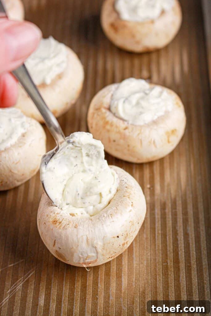 Cream Cheese Stuffed Mushrooms Recipe with Garlic and Herbs 7 Cheese mixture being added to the center of mushroom on a baking dish.