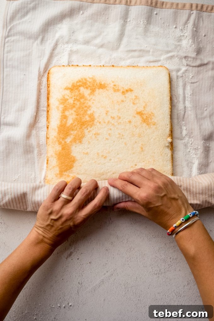 Decadent Strawberry Swiss Roll Recipe with Cream Filling 12 Someone is rolling up a layer of cake into a kitchen towel to make a Swiss roll cake.