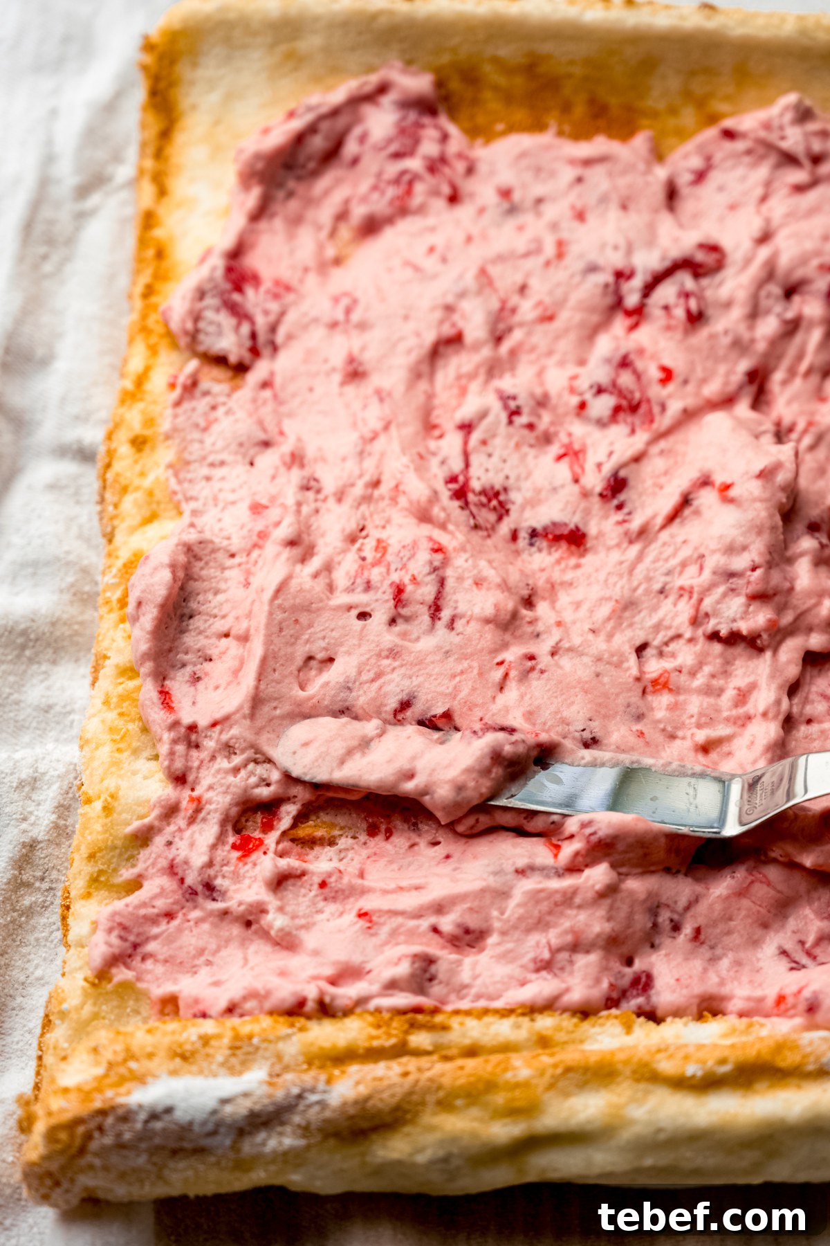 Decadent Strawberry Swiss Roll Recipe with Cream Filling 17 Someone is using a spatula to spread the filling into a Swiss roll cake.