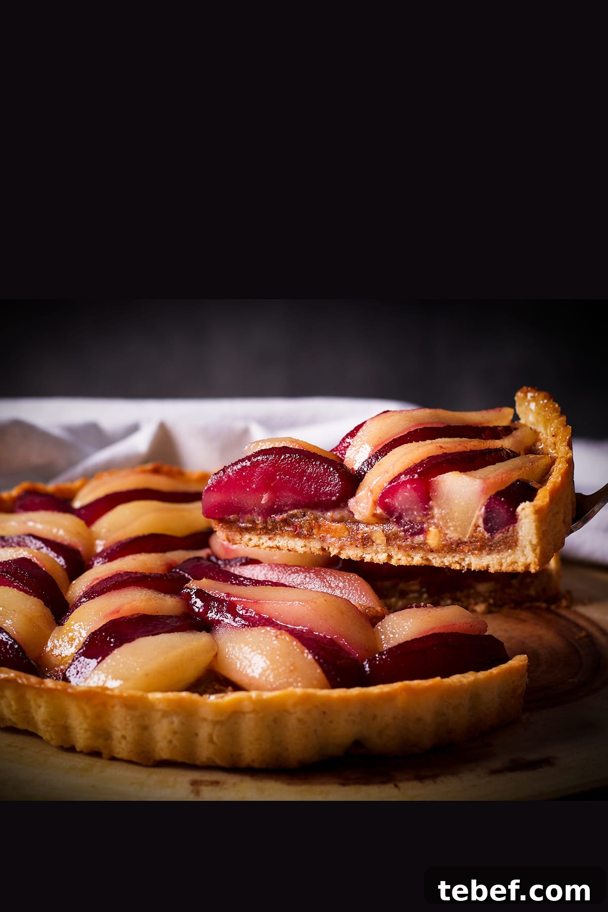 Someone using a pie server to lift a slice of poached pear and frangipane tart from a serving platter.