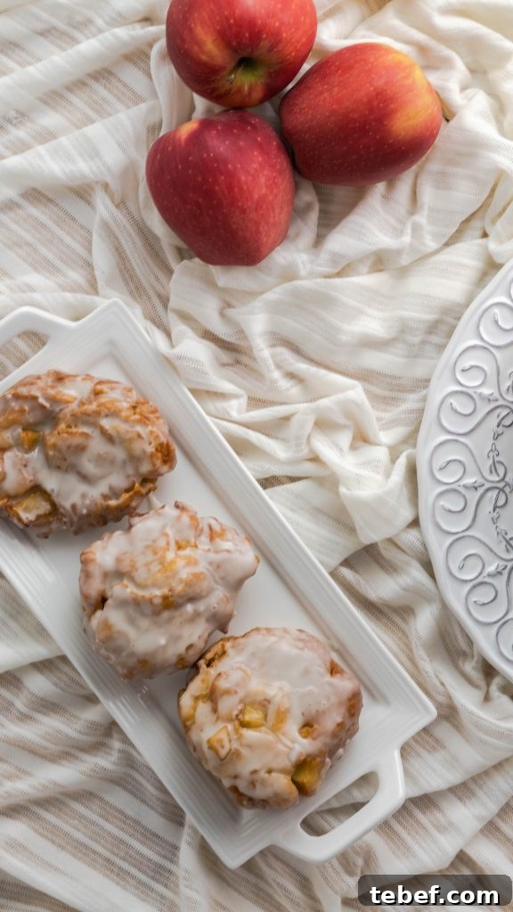 Crispy Homemade Apple Fritters Recipe for Irresistible Fall Treats 3 Glazed apple fritters on a rectangular plate with three apples next to it.