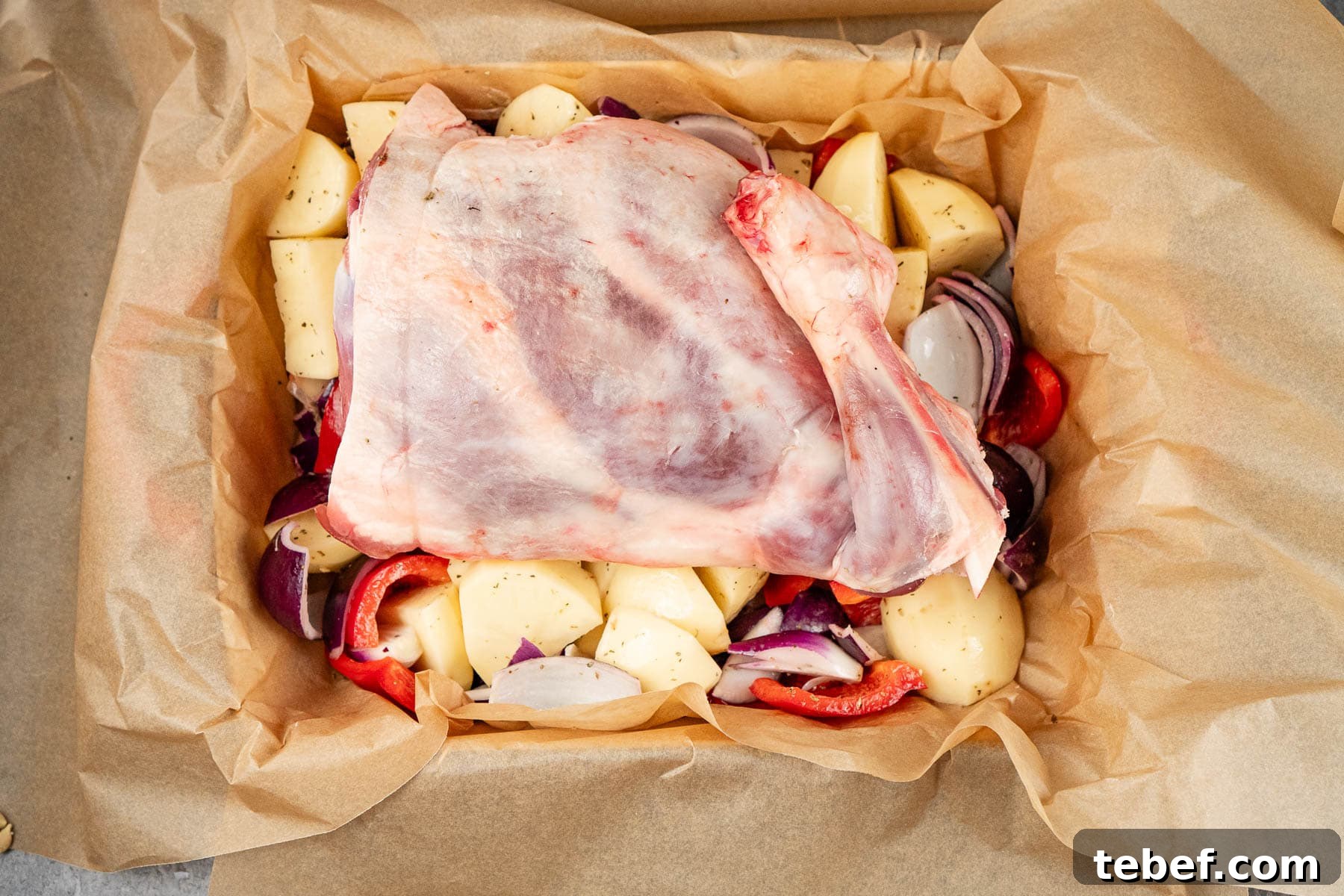 A leg of lamb on a bed of chopped potatoes,red onion and red pepper in a parchment paper lined oven dish