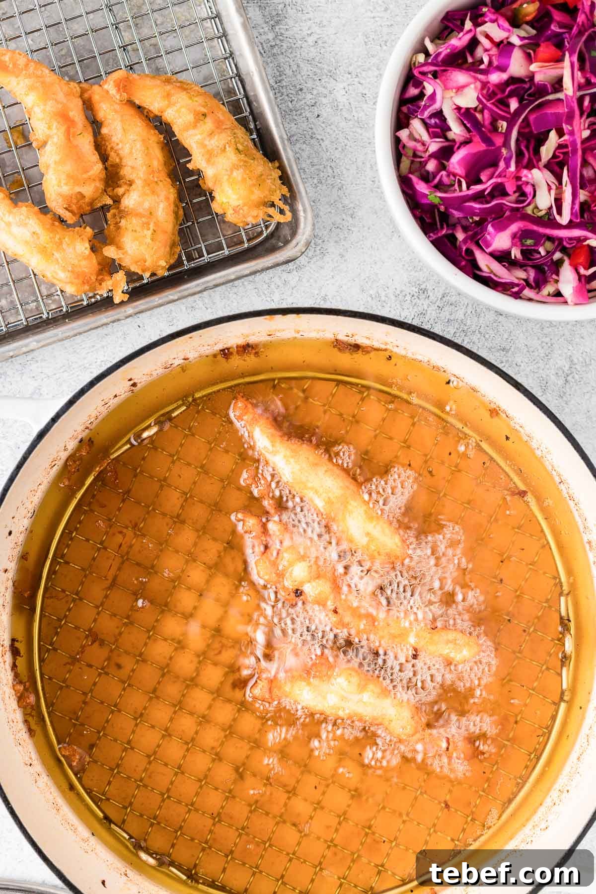 Crispy Beer-Battered Shrimp Recipe — Crunchy Fried Shrimp Guide 6 Fried beer-battered shrimp cooking in hot oil with a wire rack and bowl of sliced cabbage salad in the background.