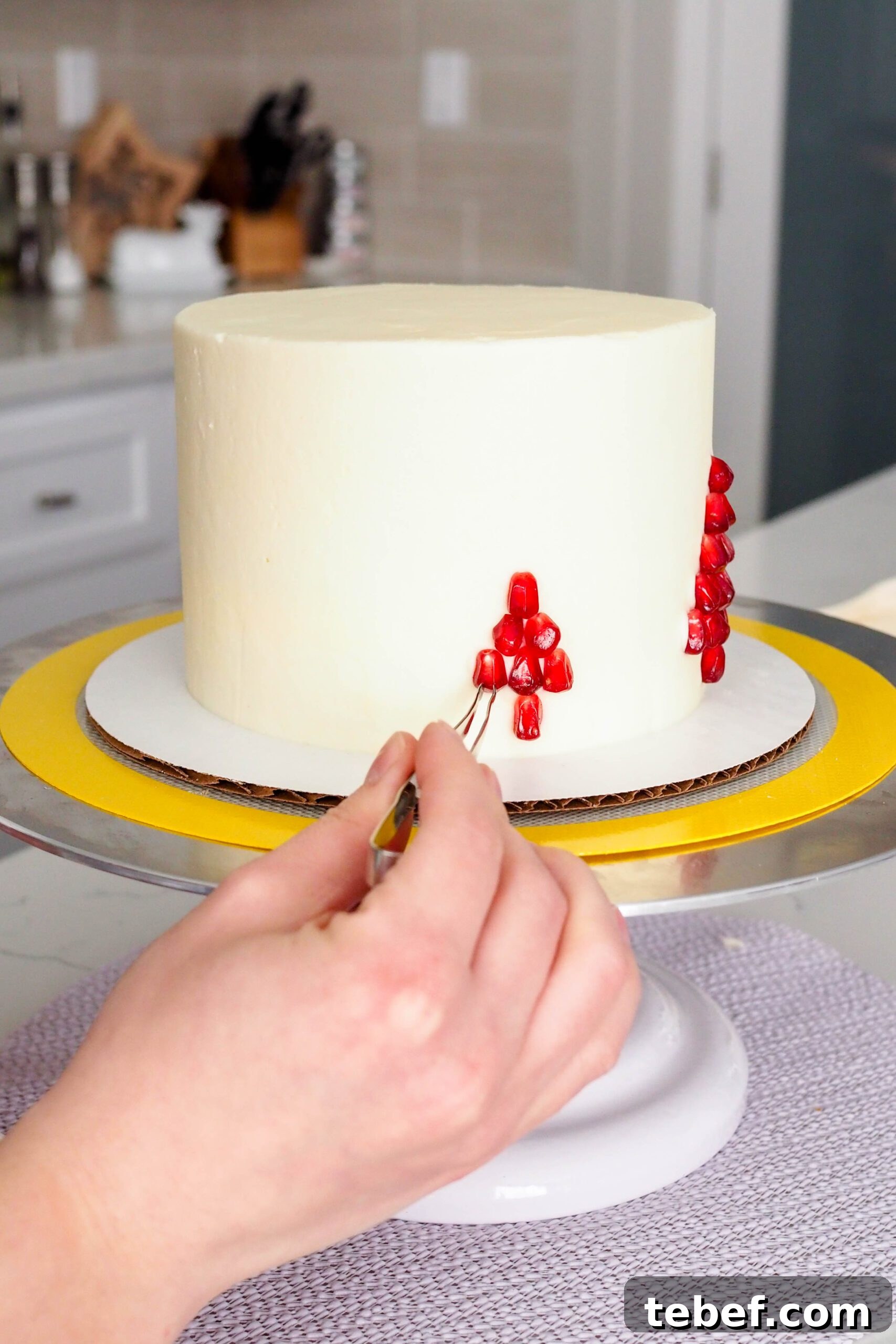White Chocolate and Pomegranate Layer Cake Recipe 15 A hand uses decorating tweezers to place pomegranate arils on the side of a cake.