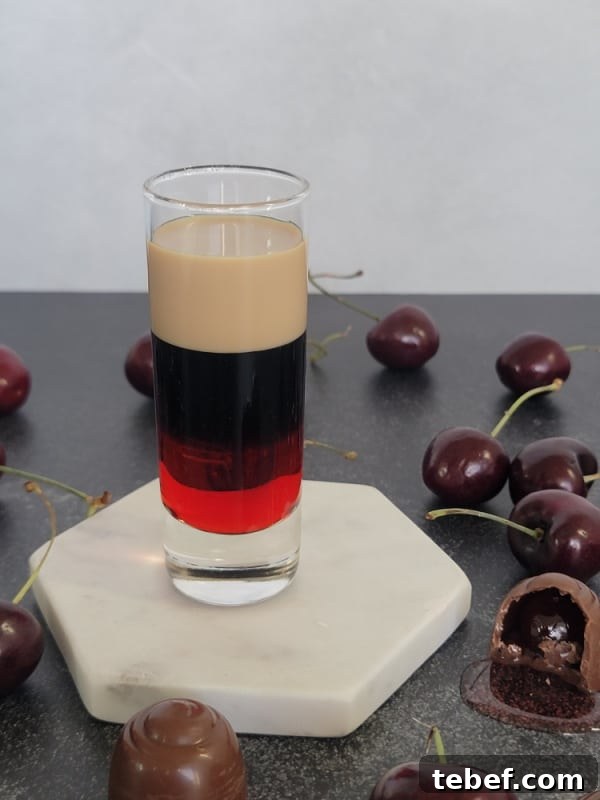 Chocolate covered cherry layered shot in a glass shot glass on a white coaster surrounded by chocolate covered cherry candies
