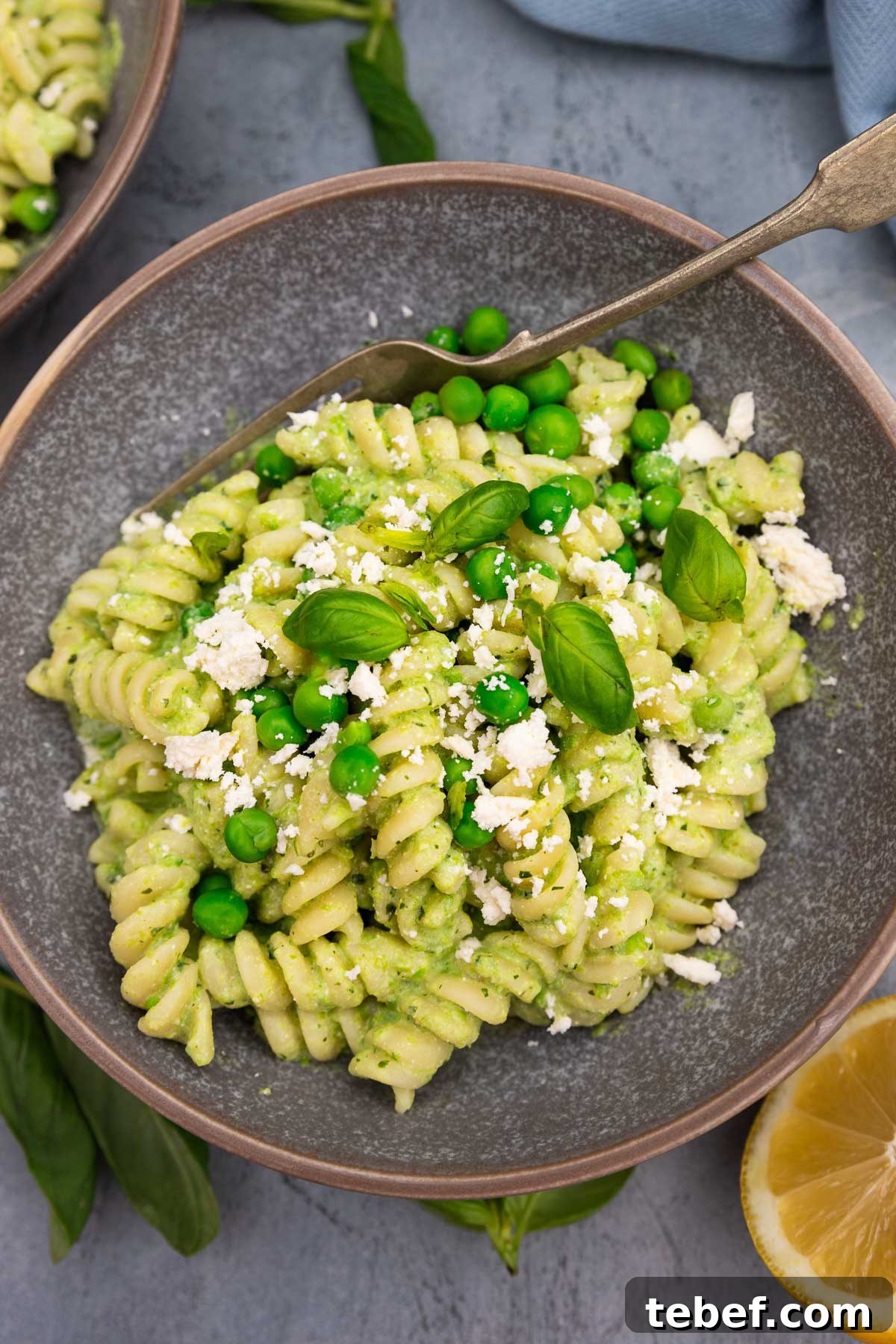 Creamy Green Pea Pasta with Frozen Peas — No Cream Needed 2 Closeup of a mottled grey bowl of fusilli pasta with peas with fresh peas and feta and herbs on top on a grey background with a fork in the bowl and lemon halves and beside it.