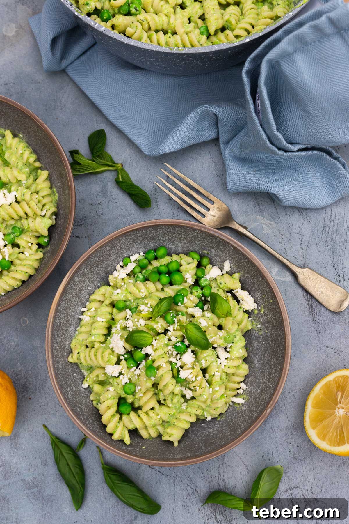 Creamy Green Pea Pasta with Frozen Peas — No Cream Needed 7 Overhead view of two bowls of creamy pea pasta with fresh peas, herbs and feta on top on a blue distressed background with more fresh herbs, lemon halves and a pale blue tea towel around them.