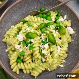 Creamy Green Pea Pasta with Frozen Peas — No Cream Needed 8 Closeup of a mottled grey bowl of fusilli pasta with peas with fresh peas and feta and herbs on top on a grey background with a fork in the bowl and lemon halves and beside it.