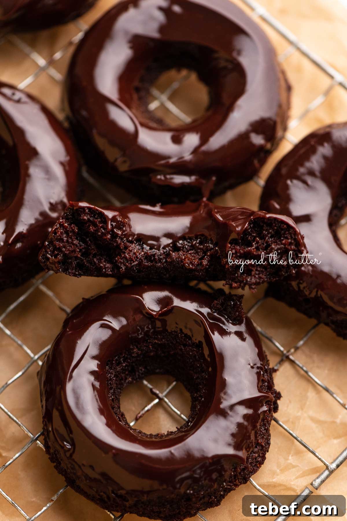 Homemade Baked Chocolate Donuts Recipe for Moist, Rich Donuts 3 Half of a homemade baked chocolate donut topped with chocolate ganache on a wire cooling rack with more donuts | © Beyond the Butter®