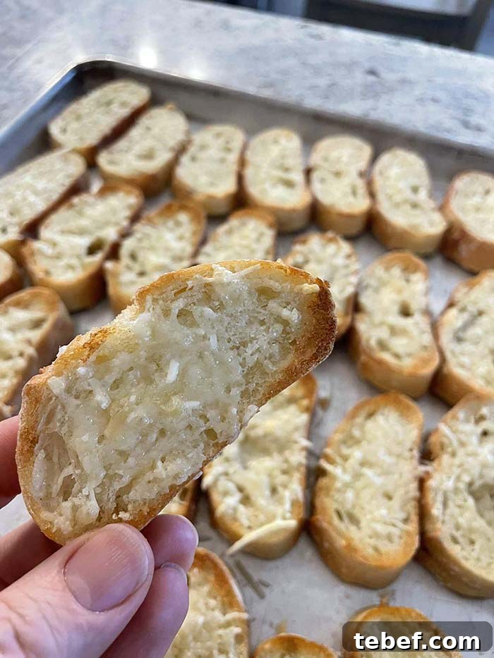 A toasted parmesan baguette slice held above a baking sheet full of sliced toasted parmesan baguette pieces.