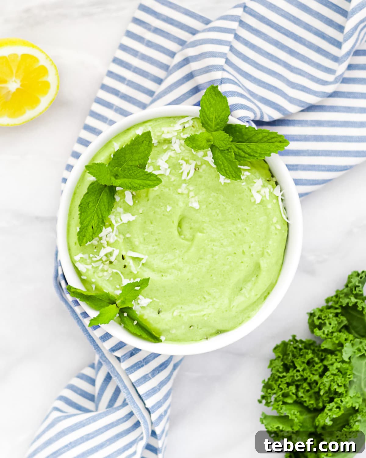A smoothie in a white bowl, garnished with coconut flakes and 3 fresh sprigs of mint. Sitting on a blue and white stripped cloth, with lemon and kale in the background.