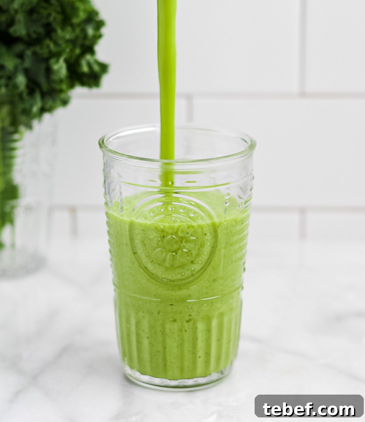 A green smoothie being poured into a clear glass. A bunch of kale is in the background.