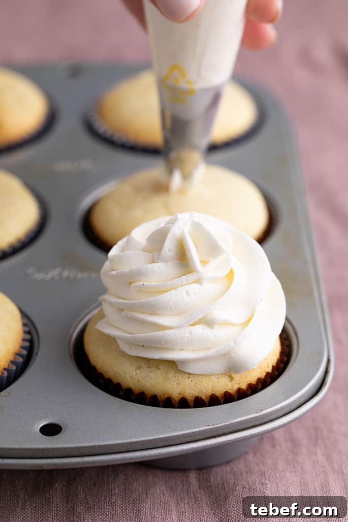 Gluten-Free Vanilla Cupcakes Recipe for Moist, Fluffy Results 16 6 baked vanilla cupcakes in a muffin tin one with frosting swirl and hand holding piping bag on top of second cupcake in background