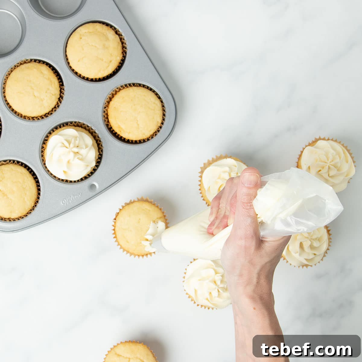 Gluten-Free Vanilla Cupcakes Recipe for Moist, Fluffy Results 23 hand holding clear pastry bag with white frosting squeezing from a metal piping tip onto a yellow vanilla cupcake