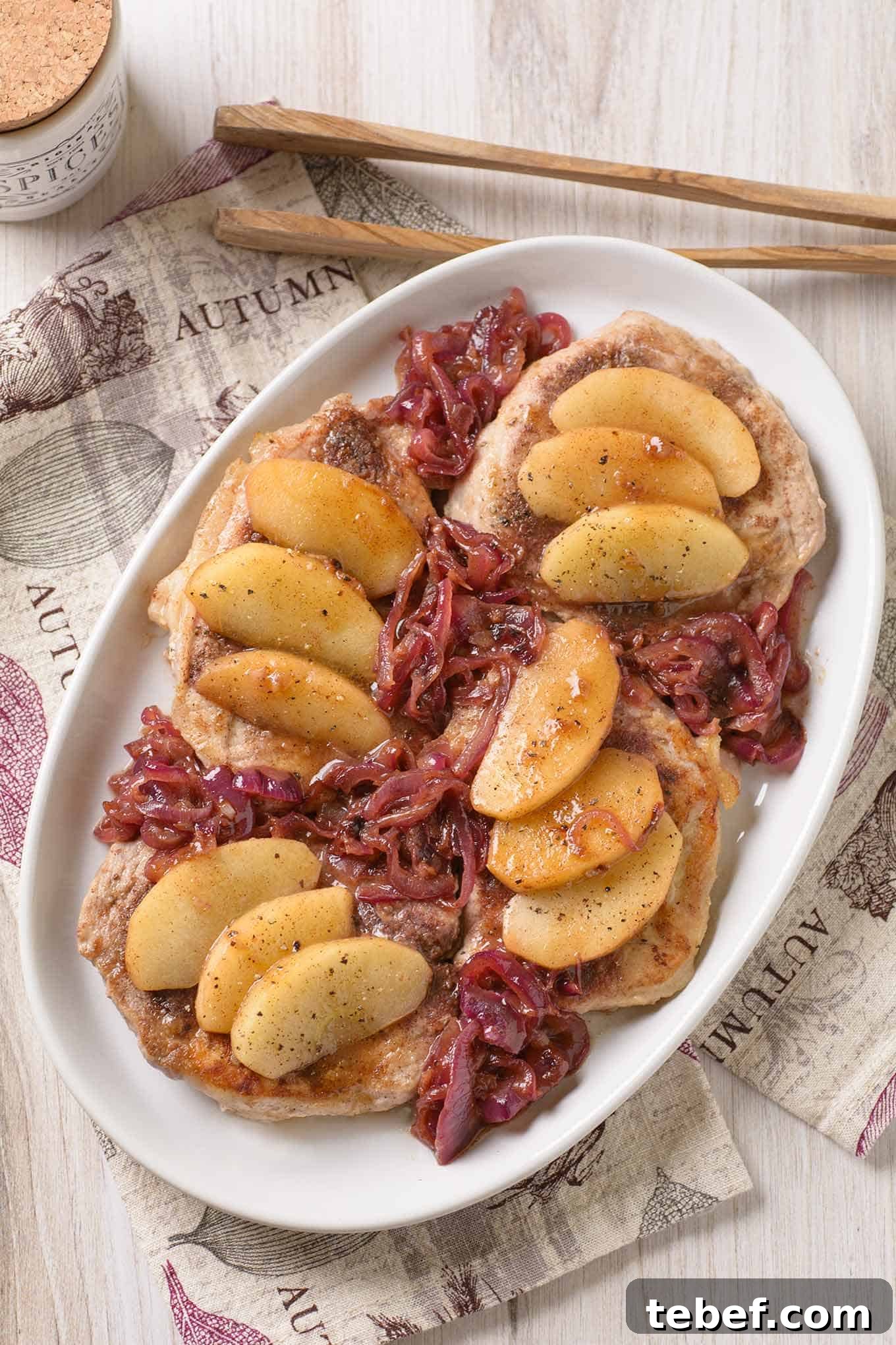 Spiced Pork Chops with Caramelized Apples and Onions 2 A an overhead view of boneless pork chops with apple slices and caramelized red onions arranged on top - a printed tea towel and serving tongs in the background.