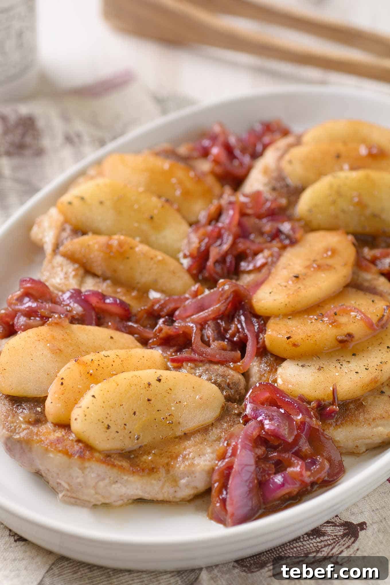 Spiced Pork Chops with Caramelized Apples and Onions 3 A close view of a boneless pork chops topped with apple slices, cider pan sauce, and caramelized red onions on a serving platter,