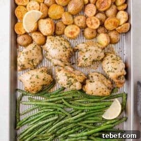 Sheet pan with roasted chicken, halved potatoes, and green beans, garnished with lemon wedges.
