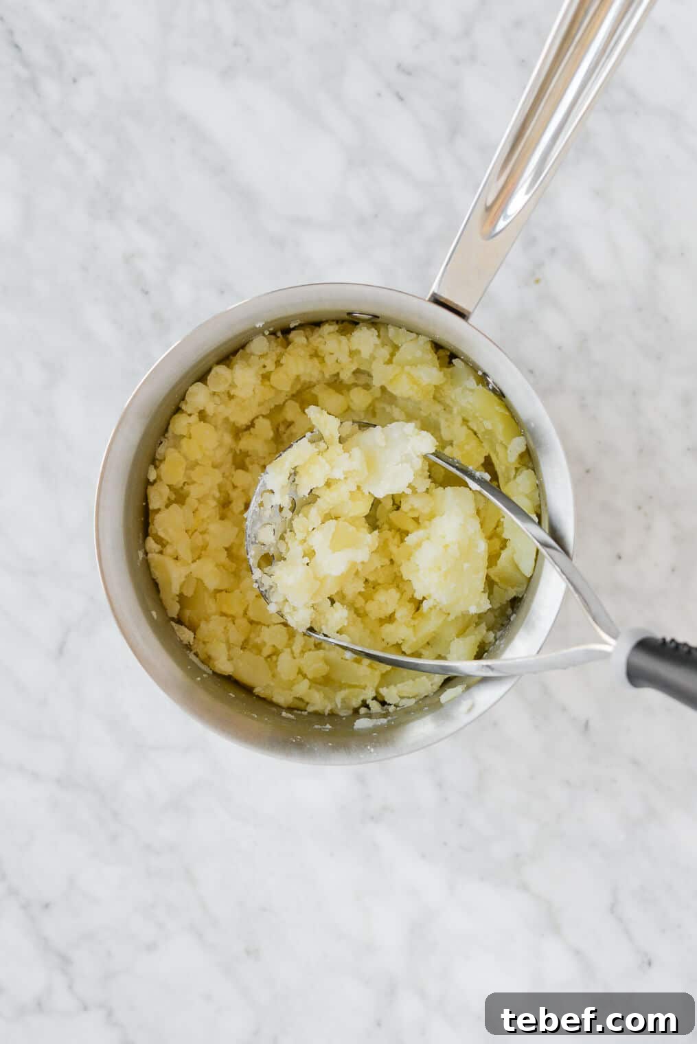 Ultra-Creamy Mashed Potatoes Recipe for Perfect Texture 6 potatoes being mashed with a potato masher in a pot sitting on a marble surface