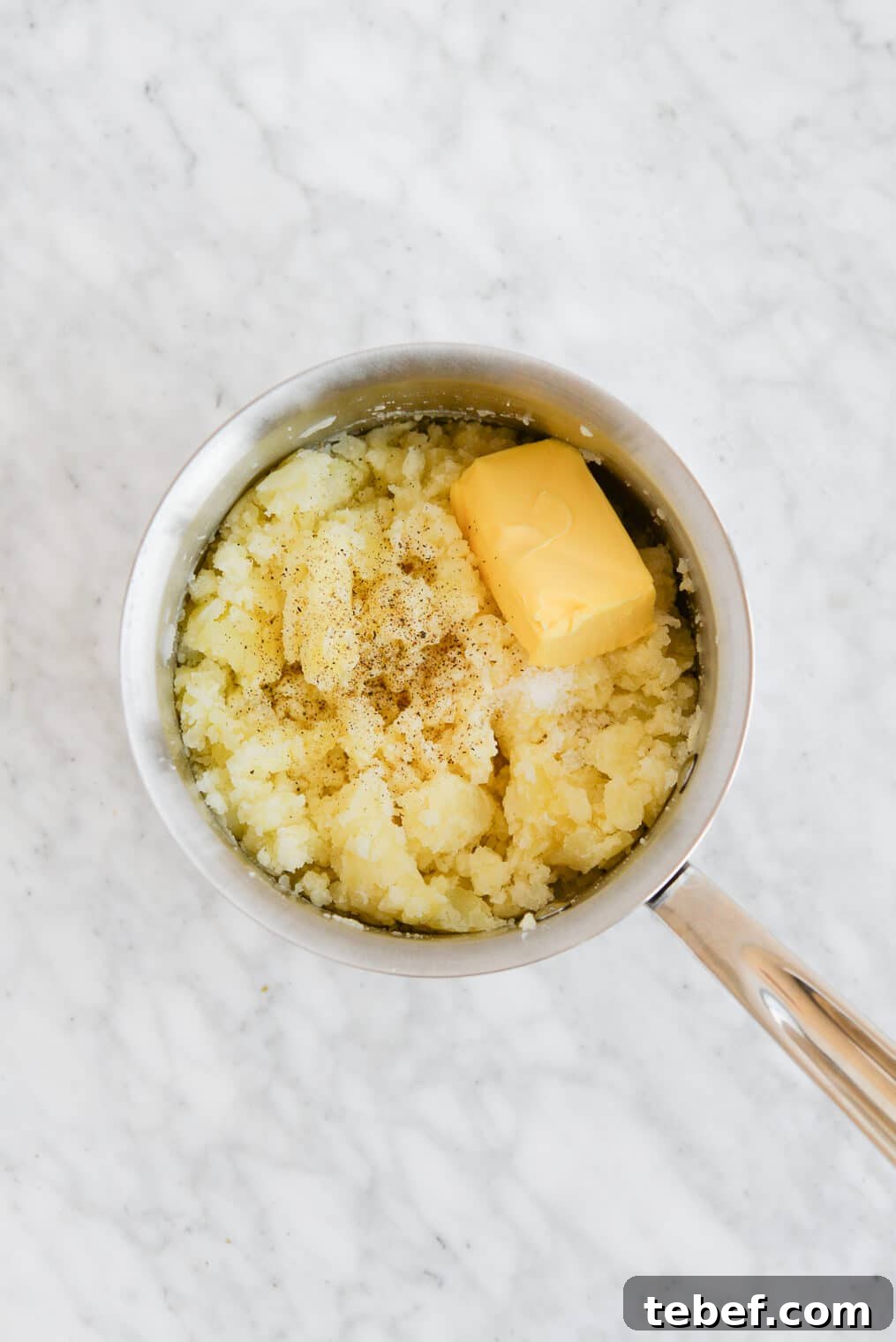 Ultra-Creamy Mashed Potatoes Recipe for Perfect Texture 7 top view of a pot of mashed potatoes with a stick of butter melting on top of the mashed potatoes sitting on a marble surface