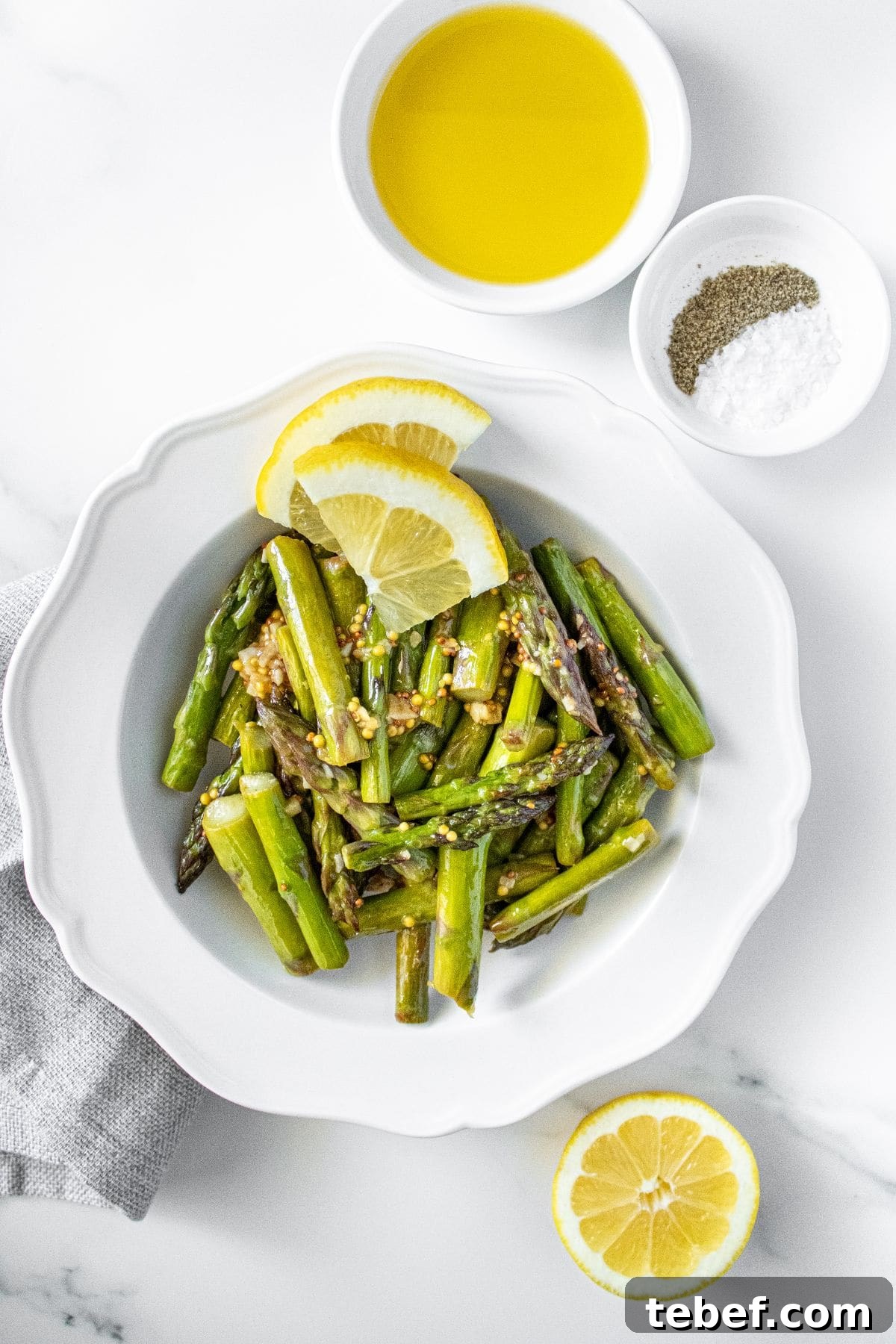 Crispy Garlic Sautéed Asparagus Recipe 5 White dish with Sautéed Asparagus, garnished with some lemon slices, with some pots of seasoning around the edge.