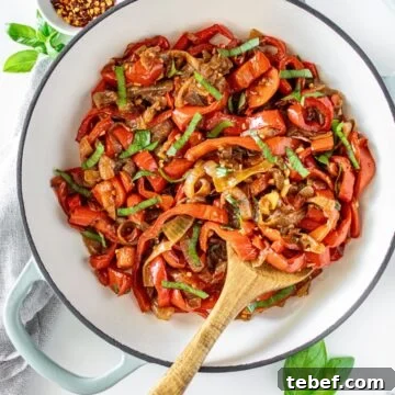 Sautéed Peppers and Onions in a large ceramic dish with a wooden spoon, and some basil leaves and chilli around the edge.