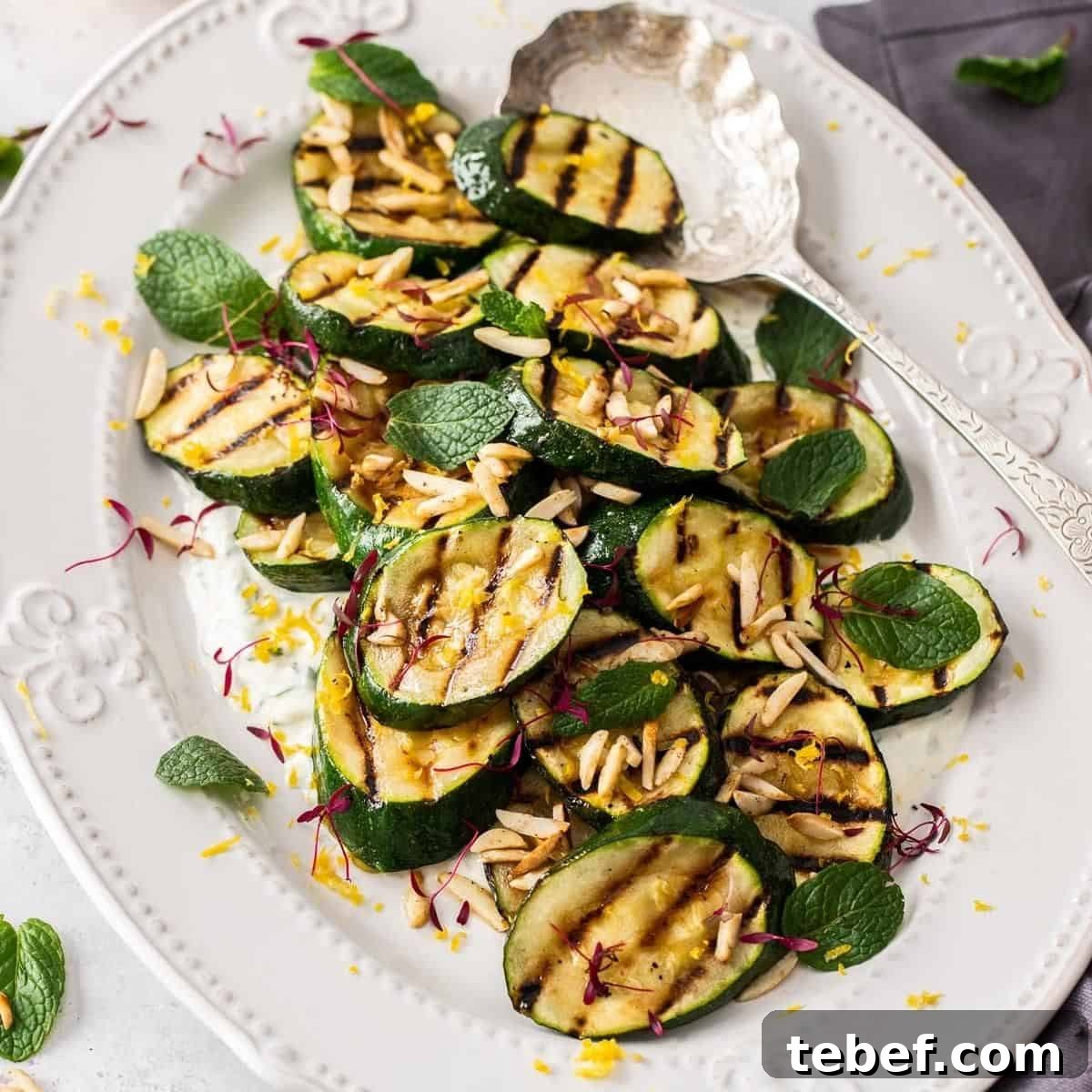 Oval white platter with grilled zucchini, garnished with slivered almonds, with a silver serving spoon on edge of the plate.