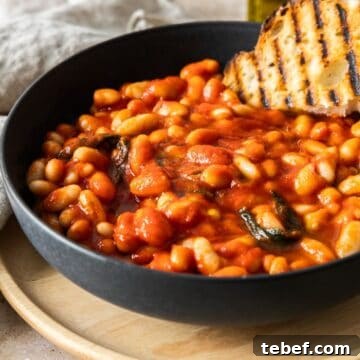 A serving of Tuscan Stewed Beans in a round black bowl, with a piece of grilled bread sitting on the beans.