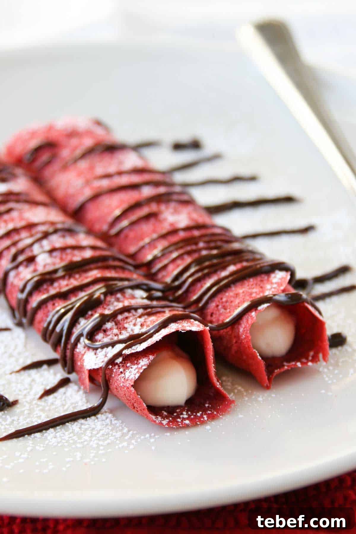 Red velvet crepes are made from scratch and then filled with a simple cheesecake filling. A chocolate drizzle finishes it for a decadent finish. Two rolled red velvet crepes filled with cream cheese filling and drizzled with chocolate.