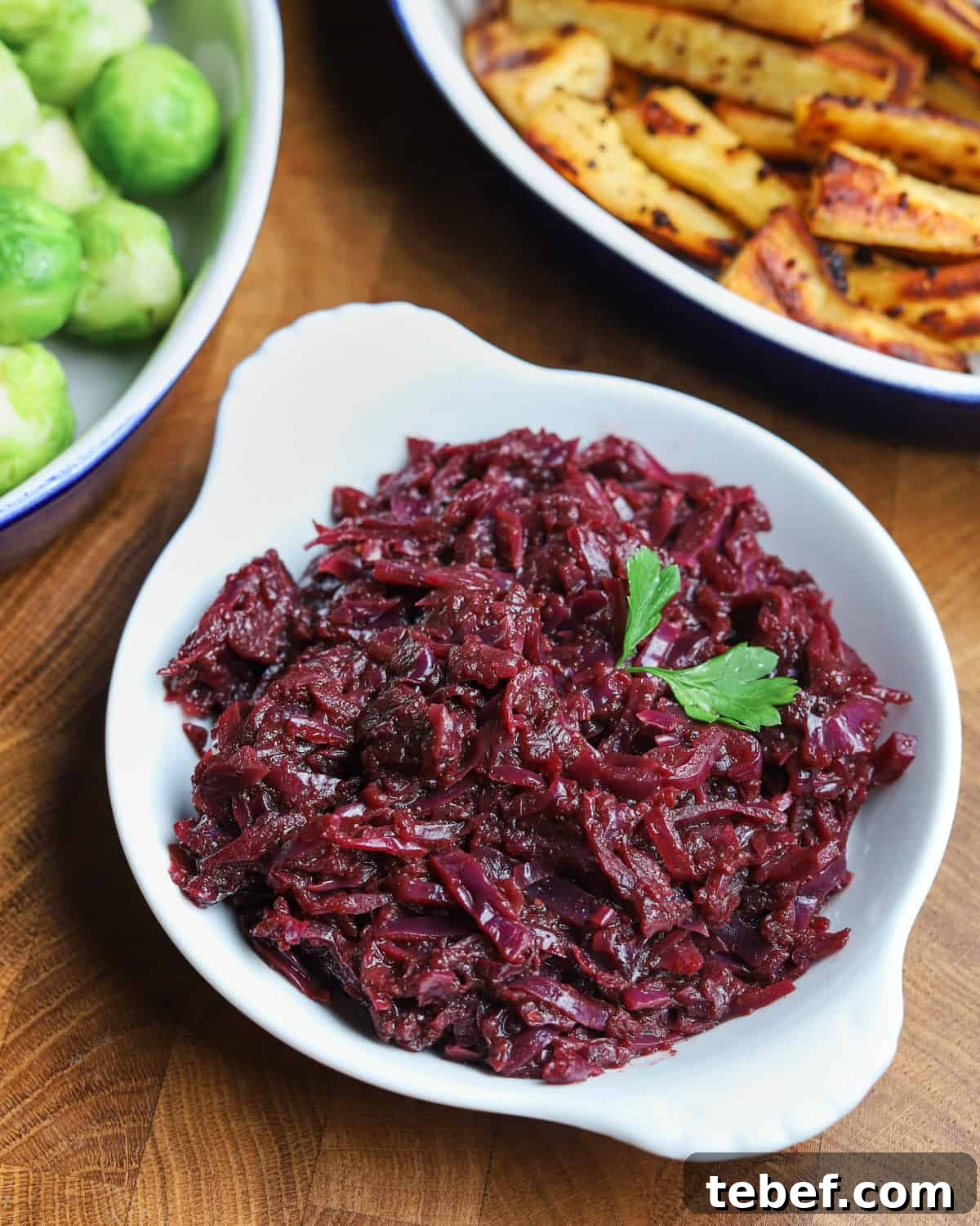 Photo of a bowl of braised red cabbage on a wooden table with a dish of sprouts and a dish of parsnips in the background.
