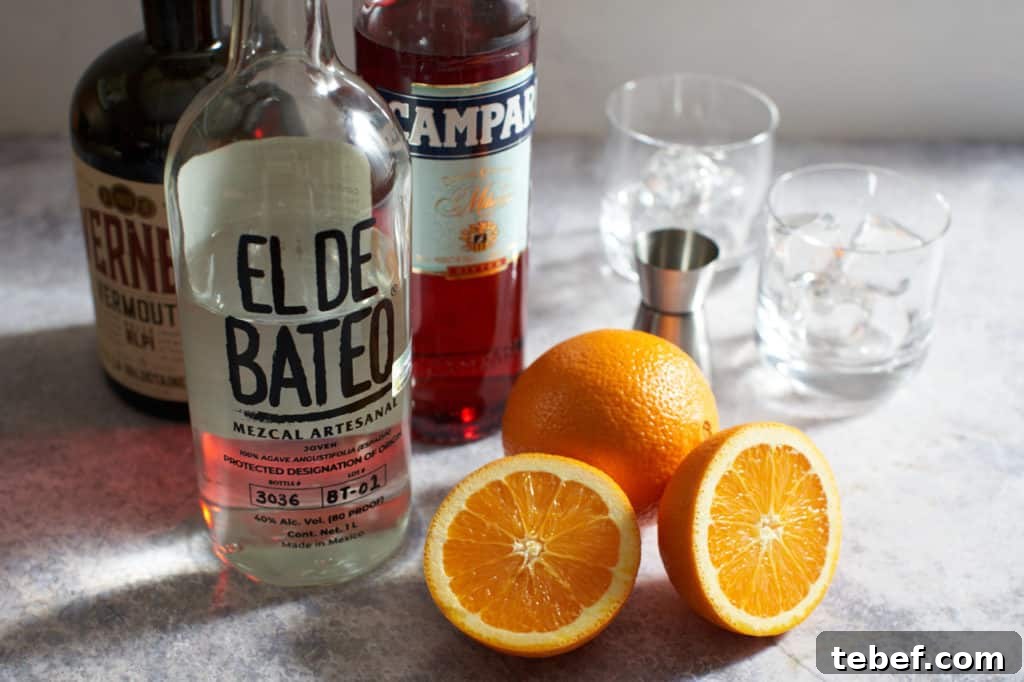 Bottles of mezcal, Campari and sweet vermouth, next to sliced oranges, a cocktail jigger and two glasses of ice.