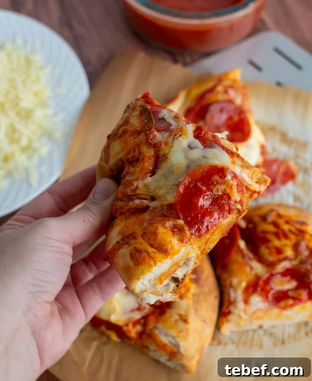 How to Bake Frozen Pizza for Crispy, Restaurant-Style Results 2 A hand holds a slice of pepperoni pizza above a counter where pizza slices, cheese, and sauce are visible.