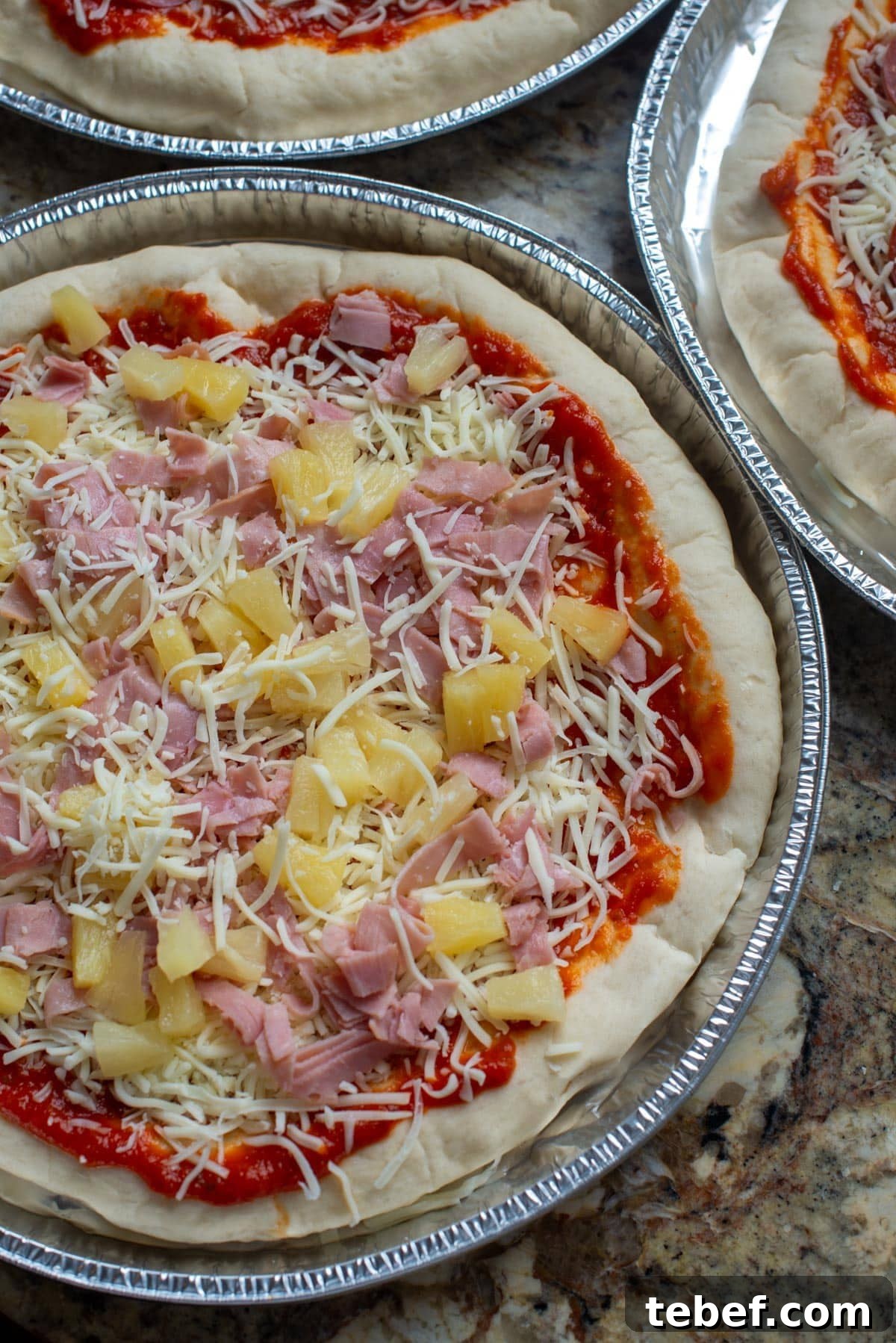 How to Bake Frozen Pizza for Crispy, Restaurant-Style Results 3 Prepared pizzas topped with sauce, cheese, ham, and pineapple sit in disposable containers prior to being frozen.