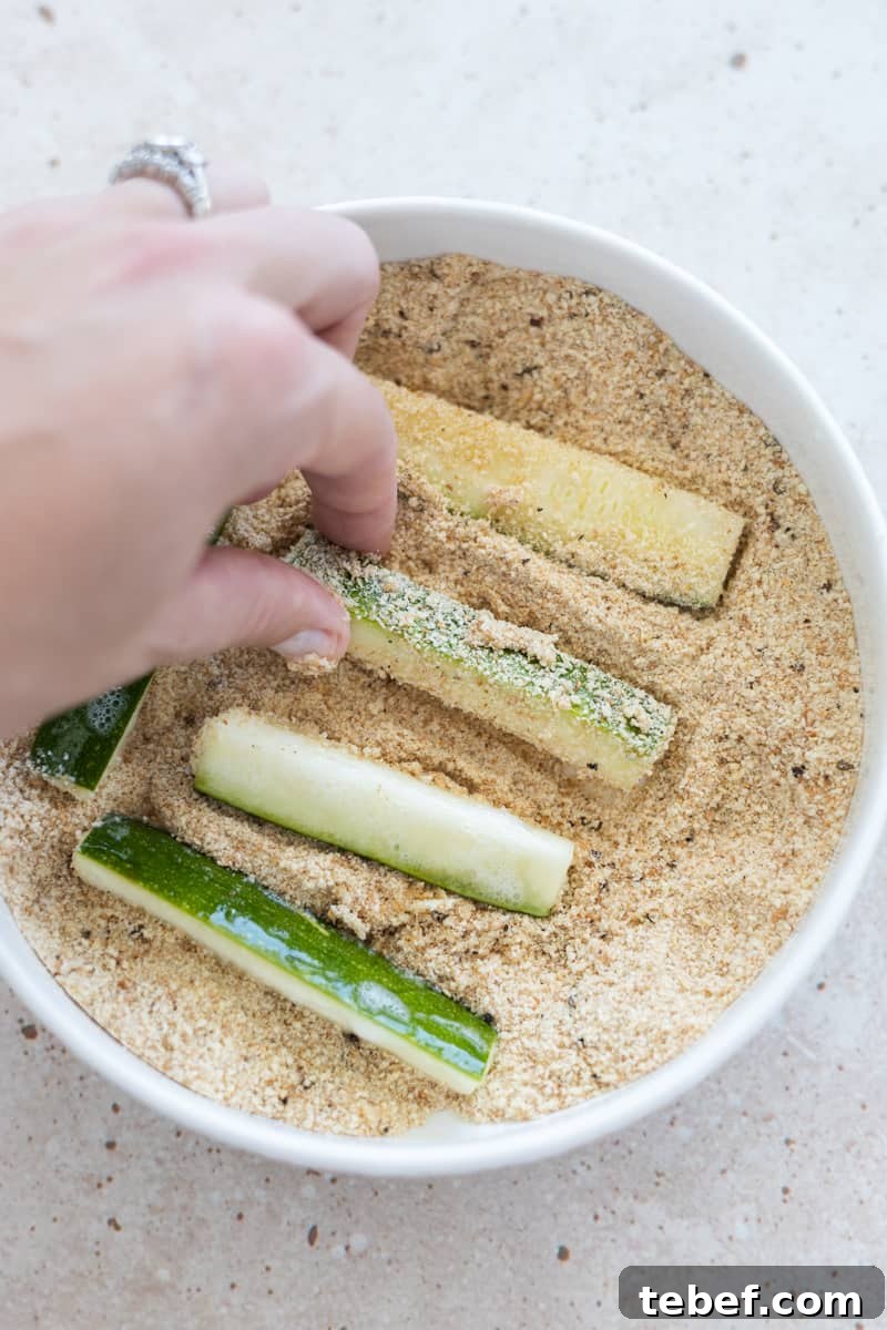 A hand tossing zucchini slices in a bowl with the breadcrumb mixture. 