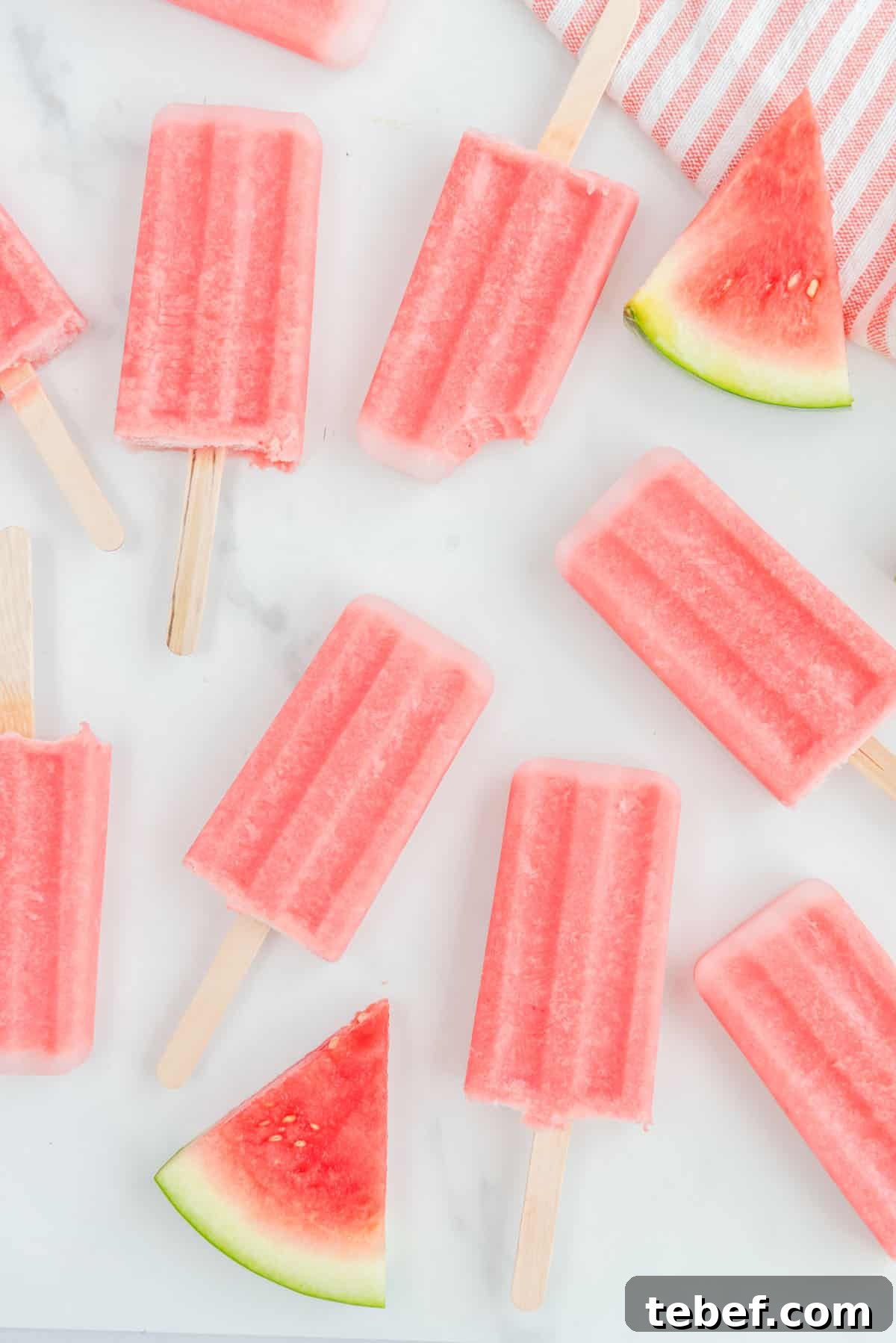 Refreshing Watermelon Popsicle Recipe: 3-Ingredient Frozen Treats 12 Overhead photo of multiple watermelon popsicles accented by watermelon slices in the upper righthand and lower lefthand corners. One of the popsicles has a small bite taken out of it.