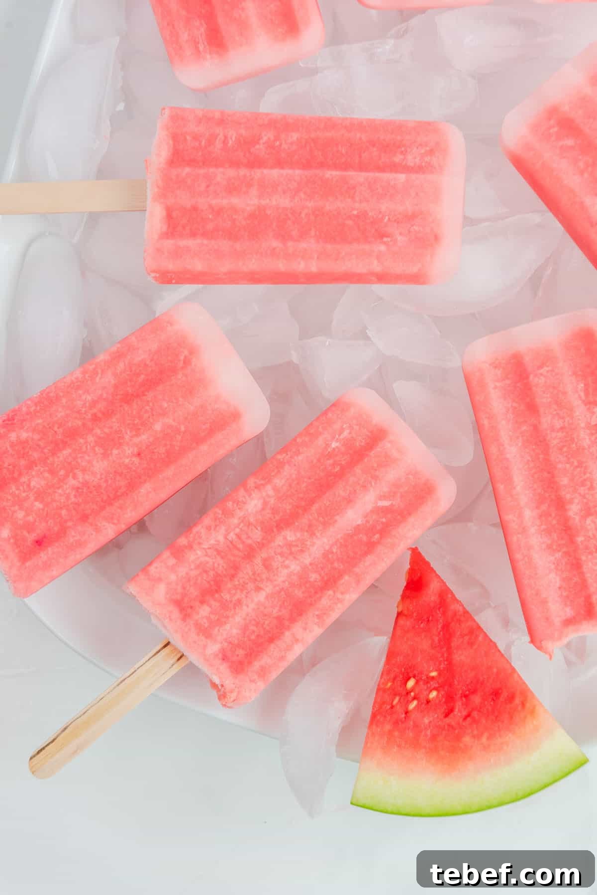 Refreshing Watermelon Popsicle Recipe: 3-Ingredient Frozen Treats 13 Close-up, overhead photo of a plate of ice cubes topped with watermelon popsicles stacked on top of each other. Accented by a sliced watermelon on the bottom of the photo.