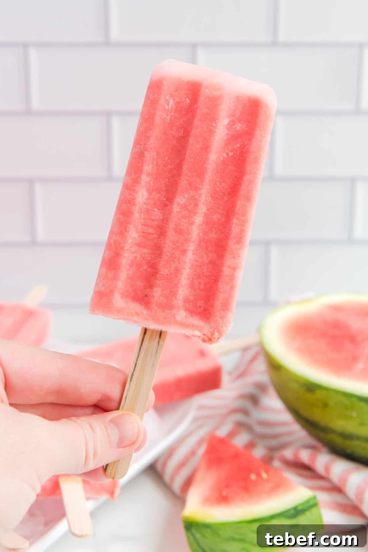 Refreshing Watermelon Popsicle Recipe: 3-Ingredient Frozen Treats 3 A hand holding a single popsicle. The background is filled with more popsicles and sliced watermelons.