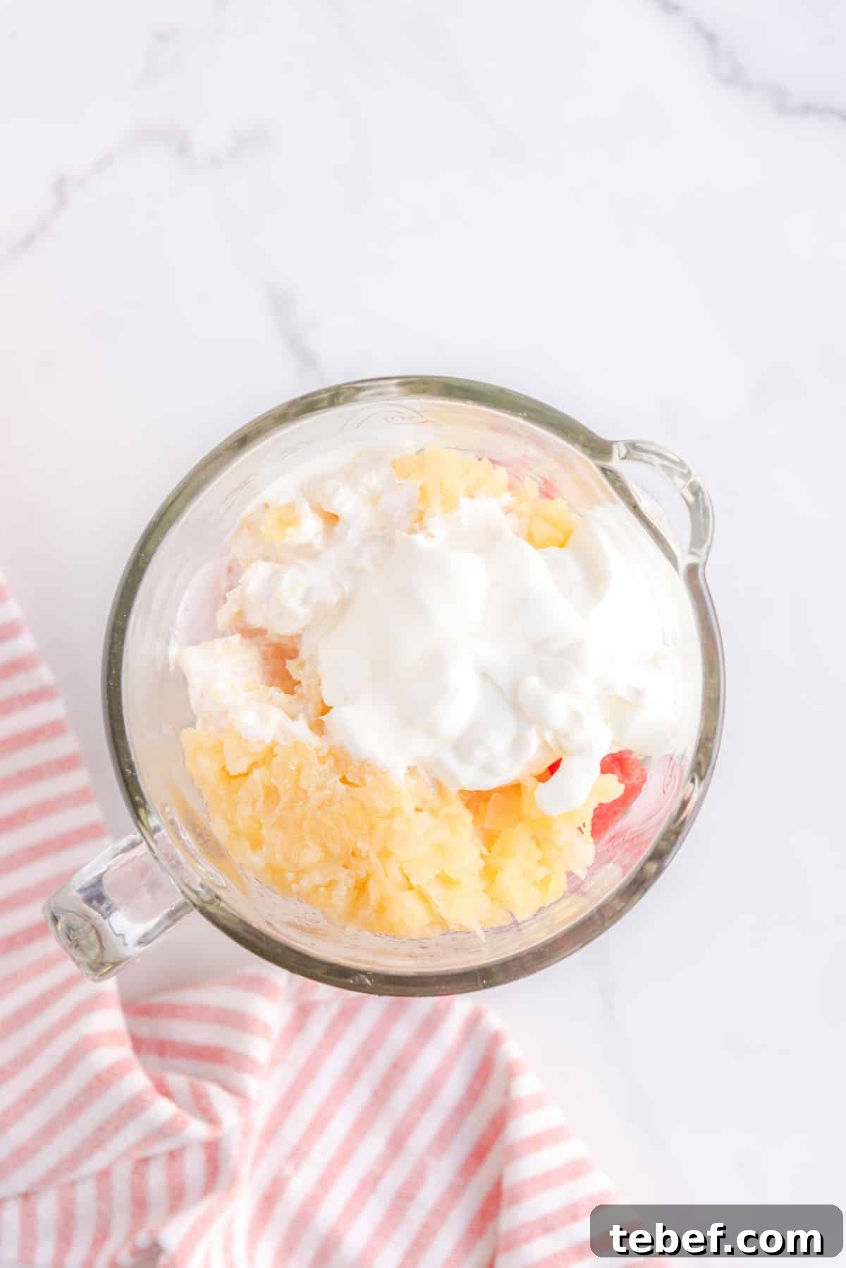 Refreshing Watermelon Popsicle Recipe: 3-Ingredient Frozen Treats 6 Overhead shot of blender filled with watermelon, crushed pineapple, heavy cream, lemon juice and greek yogurt. Bottom left corner accented with a pink and white-striped cloth.