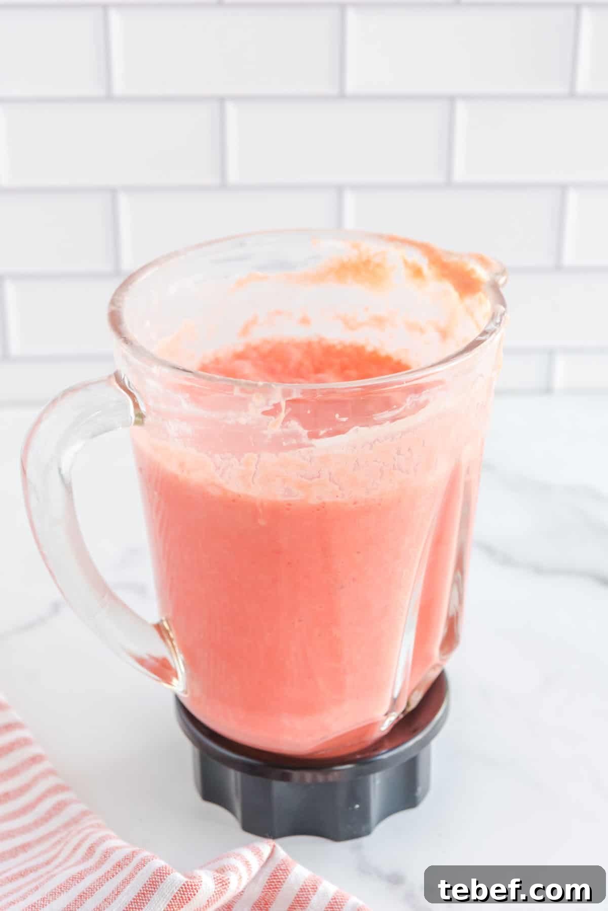 Refreshing Watermelon Popsicle Recipe: 3-Ingredient Frozen Treats 7 Straight on shot of blender with fully blended watermelon, crushed pineapple, heavy cream, lemon juice and greek yogurt. Bottom left corner accented with a pink and white-striped cloth.