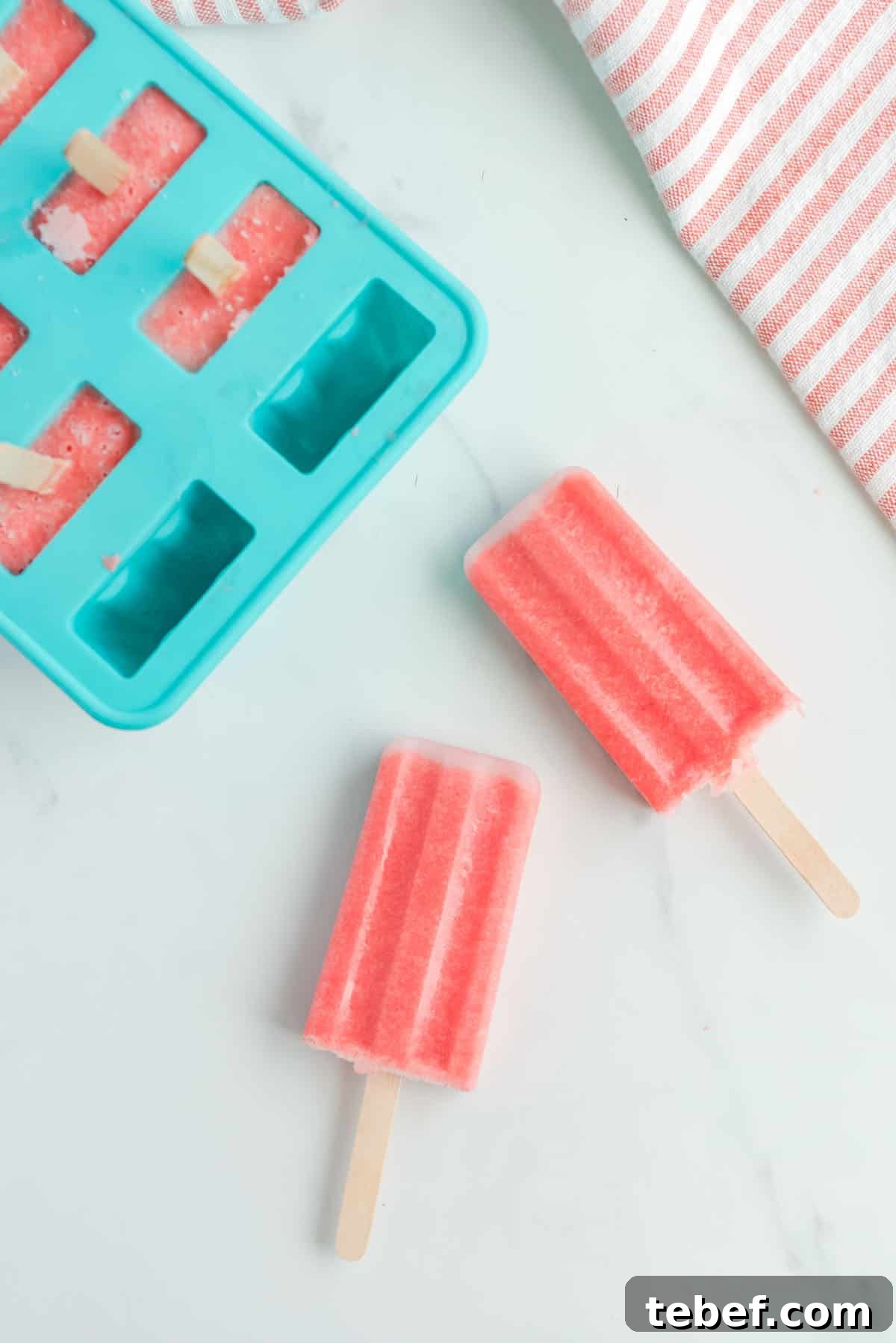 Refreshing Watermelon Popsicle Recipe: 3-Ingredient Frozen Treats 10 Two watermelon popsicles were removed from the popsicle mold. The mold is on the left hand side of the photo and the righthand side is accented by a pink and white striped cloth.