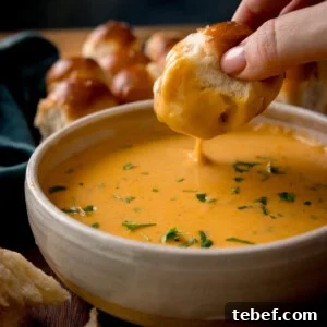 Creamy Beer Cheese Dip Recipe for Game Day and Parties 12 A hand dips a piece of bread into a bowl of creamy orange beer cheese dip, garnished with chopped herbs. More bread rolls are visible in the background on a wooden surface.